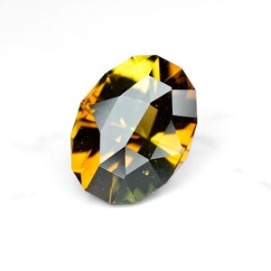 May include: A faceted gemstone with a mix of yellow, amber, and dark brown hues. The gem has a complex, multi-faceted cut, reflecting light and creating a sparkling effect. The shape is an elongated oval.