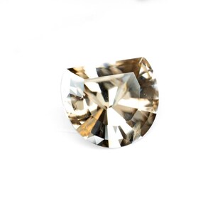 May include: A faceted, half-moon shaped gemstone with a light brown hue. The gem has multiple facets that reflect light, creating a sparkling effect. The gemstone is set against a white background.