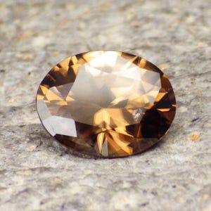 May include: A brown oval-shaped gemstone with a faceted cut. The gemstone has a translucent appearance with a slight shimmer.