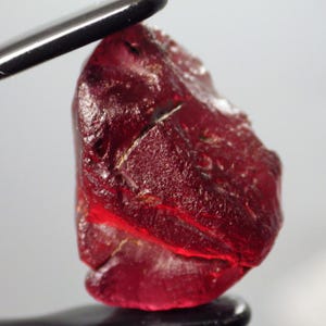 May include: A deep red, faceted gemstone with a rough, natural texture. The gemstone is a ruby, a variety of corundum.