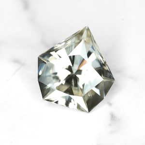 May include: A faceted, light-colored gemstone with a unique, geometric cut. The gem has a complex internal structure, reflecting light to create a brilliant sparkle. The stone is set against a white background, highlighting its clarity and shape. This gemstone is ideal for jewelry.