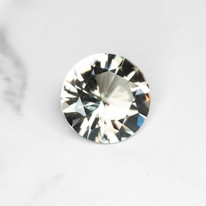 May include: A round, faceted gemstone with a brilliant cut. The gem is clear with internal reflections, set against a white marble surface. The facets create a play of light and shadow, highlighting its clarity and sparkle.