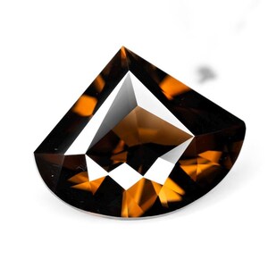 May include: A faceted gemstone with a semi-circular shape, featuring a deep amber and dark brown color palette. The stone displays sharp geometric cuts and internal light reflections that create a high-contrast pattern of dark and bright areas.