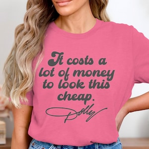 May include: A bright pink short-sleeved t-shirt featuring dark gray text that reads, It costs a lot of money to look this cheap. followed by the signature Dolly in a cursive script font centered on the front of the garment.