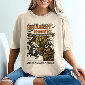 Può includere: Una t-shirt color crema con una stampa grafica che riporta il testo HELLBENT HONEYS, THE WITCHIEST SHOW ON EARTH, LIVE IN SALEM e OCT 31. Il design include tre streghe, pipistrelli, zucche e dettagli su un rodeo e gare di scopa.