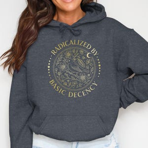 May include: A dark heather grey hooded sweatshirt featuring a gold-coloured circular graphic on the chest. The design includes a bird surrounded by flowers, leaves, and a crescent moon, with the text "RADICALIZED BY BASIC DECENCY" printed around it.