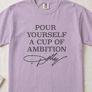 Lavender t-shirt with "POUR YOURSELF A CUP OF AMBITION Dolly" text.