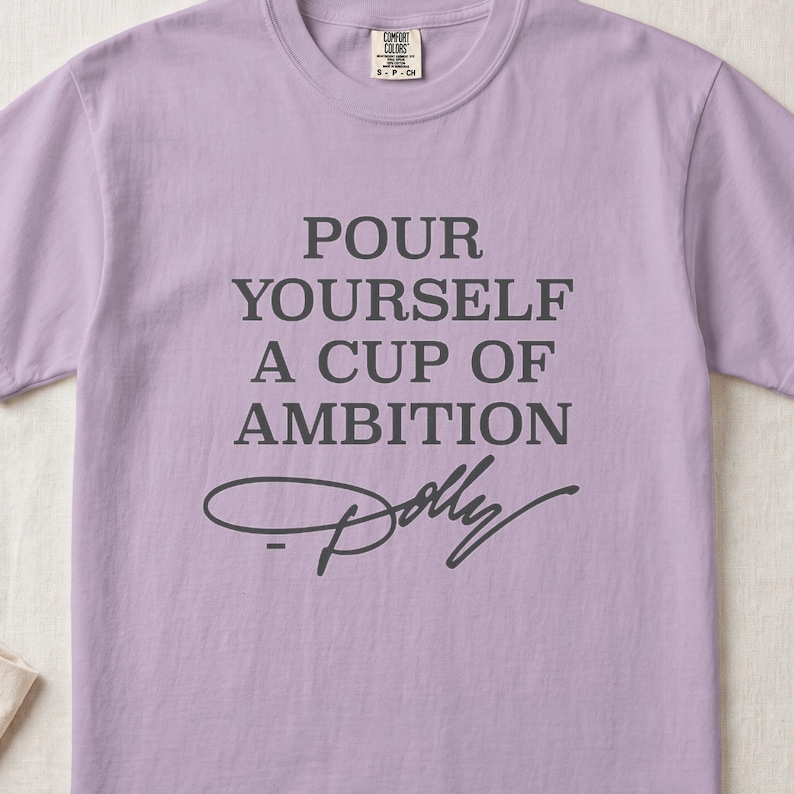 Lavender t-shirt with "POUR YOURSELF A CUP OF AMBITION Dolly" text.