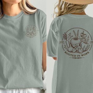Può includere: Una t-shirt a maniche corte color verde salvia con una piccola grafica circolare sul petto sinistro e una grande grafica abbinata sul retro. Il disegno raffigura varie verdure con il testo "EST. 1983" e "ROOTED IN WORK" in inchiostro marrone scuro.