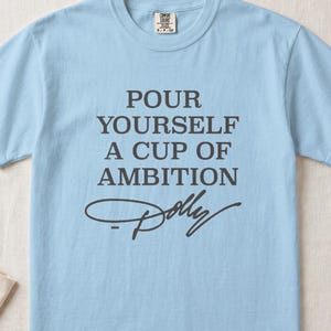 Light blue t-shirt with gray text "POUR YOURSELF A CUP OF AMBITION Dolly".