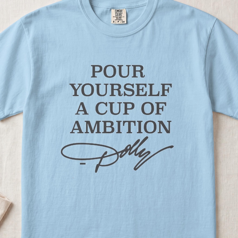 Light blue t-shirt with gray text "POUR YOURSELF A CUP OF AMBITION Dolly".