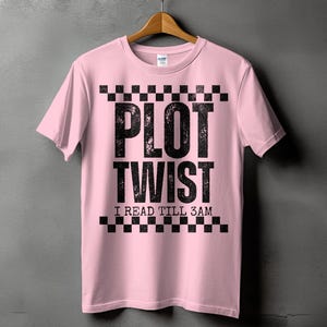 May include: A light pink short-sleeve t-shirt featuring a black checkered border at the top and bottom of the graphic. The center text reads PLOT TWIST in large distressed black letters, followed by the smaller text I READ TILL 3AM in black.