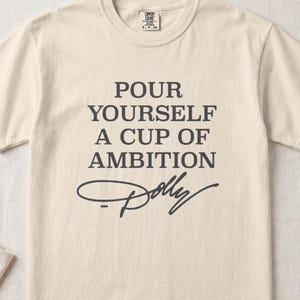 Cream t-shirt with text "Pour Yourself A Cup of Ambition" and "Dolly" signature.