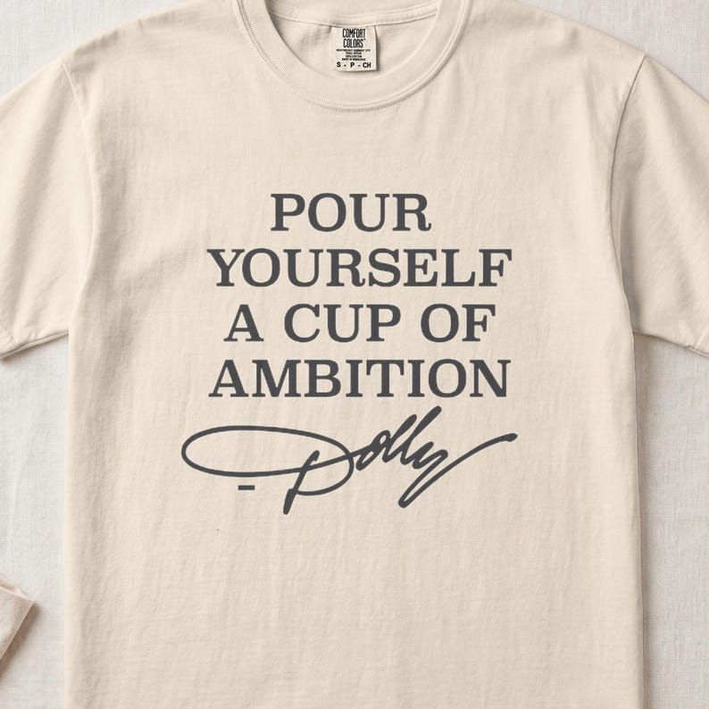 Cream t-shirt with text "Pour Yourself A Cup of Ambition" and "Dolly" signature.