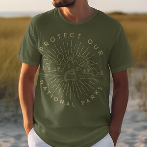May include: An olive green short-sleeve t-shirt featuring a light-colored line art graphic of mountains, a sun, clouds, trees, and a camper van. The text "PROTECT OUR NATIONAL PARKS" is printed in a circular arrangement around the central illustration.