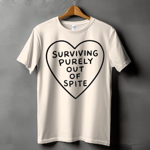 May include: A cream-coloured short-sleeve t-shirt featuring a large black heart outline on the chest. Inside the heart, the text reads "SURVIVING PURELY OUT OF SPITE" in bold, black, sans-serif capital letters. The shirt is displayed on a wooden hanger.
