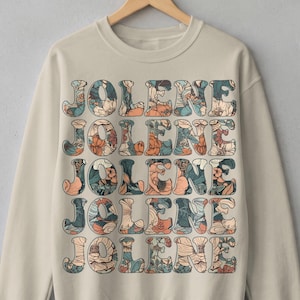 Peut inclure: Un sweat-shirt beige à col rond arborant le nom JOLENE répété cinq fois dans une grille empilée. Chaque lettre est remplie d'une illustration florale détaillée dans des tons orange sourd, bleu sarcelle, vert foncé et crème sur fond clair.