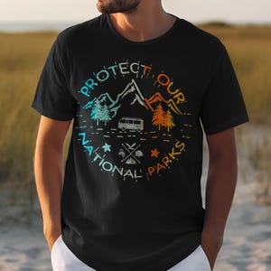 May include: A black t-shirt featuring a circular graphic with the text "PROTECT OUR NATIONAL PARKS". The design includes mountain silhouettes, pine trees, a camper van, and small icons of a fish, a tent, and a campfire in a gradient color scheme.
