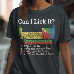 Peut inclure: Un t-shirt gris anthracite à manches courtes avec le texte Can I Lick It? au-dessus d'un tableau périodique coloré. Sous le tableau, une légende explique les niveaux de sécurité codés par couleur : Sure, Go For It, Maybe, Not The Best Idea, Please, Don't Do That, et See You On The Other Side.