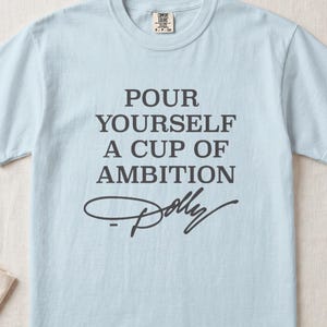 Light blue t-shirt with "Pour Yourself A Cup Of Ambition" and "Dolly" signature.