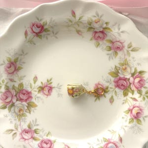 May include: A small, floral-patterned teacup charm with a gold-colored clasp rests on a white plate. The plate is decorated with a pink rose and green leaf pattern. A pink ribbon is tied in a bow above the plate.