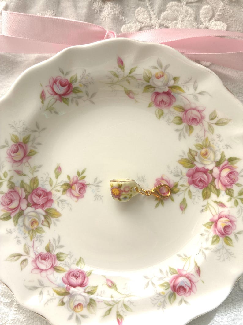 May include: A small, floral-patterned teacup charm with a gold-colored clasp rests on a white plate. The plate is decorated with a pink rose and green leaf pattern. A pink ribbon is tied in a bow above the plate.