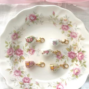 May include: A white porcelain plate with a pink rose pattern holds six miniature teacup charms. Each charm is a tiny teacup with a different floral design and a gold-colored clasp. A pink ribbon is tied in a bow at the top.