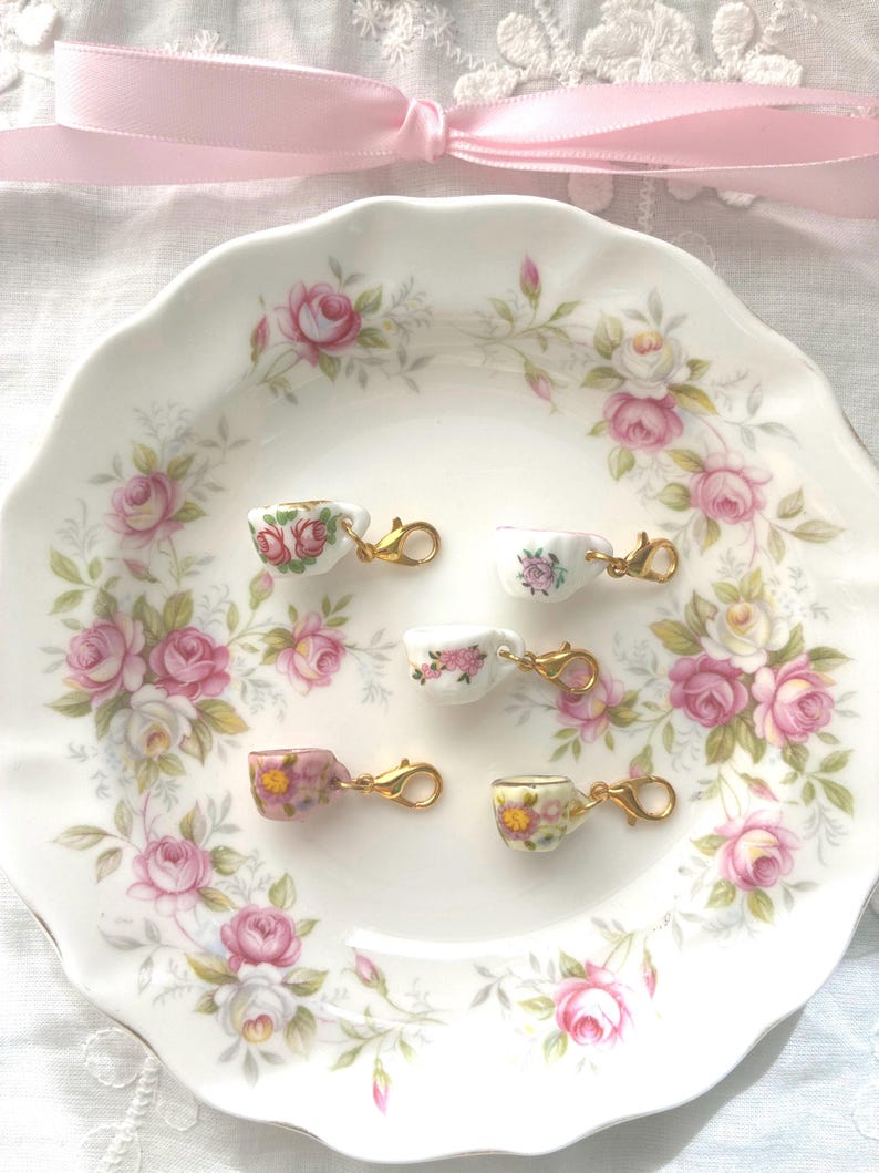 May include: A white porcelain plate with a pink rose pattern holds six miniature teacup charms. Each charm is a tiny teacup with a different floral design and a gold-colored clasp. A pink ribbon is tied in a bow at the top.