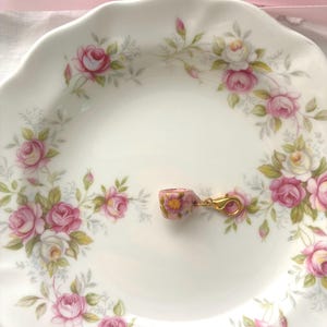 May include: A miniature pink teacup charm with a gold clasp rests on a white plate decorated with pink roses and green leaves. A pink ribbon is in the background. The teacup has a floral pattern.