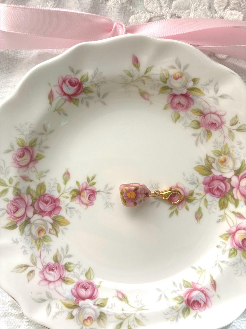 May include: A miniature pink teacup charm with a gold clasp rests on a white plate decorated with pink roses and green leaves. A pink ribbon is in the background. The teacup has a floral pattern.
