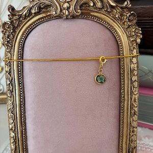 May include: An ornate gold picture frame with a pink velvet backing. A delicate gold cord is strung across the frame, holding a small, round charm with a blue floral design. The frame has detailed carvings.