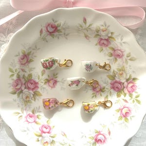 May include: A white ceramic plate with a floral rose pattern holds five miniature teacups with gold-colored clasps. Each teacup features a different floral design. A pink ribbon is tied in a bow above the plate.