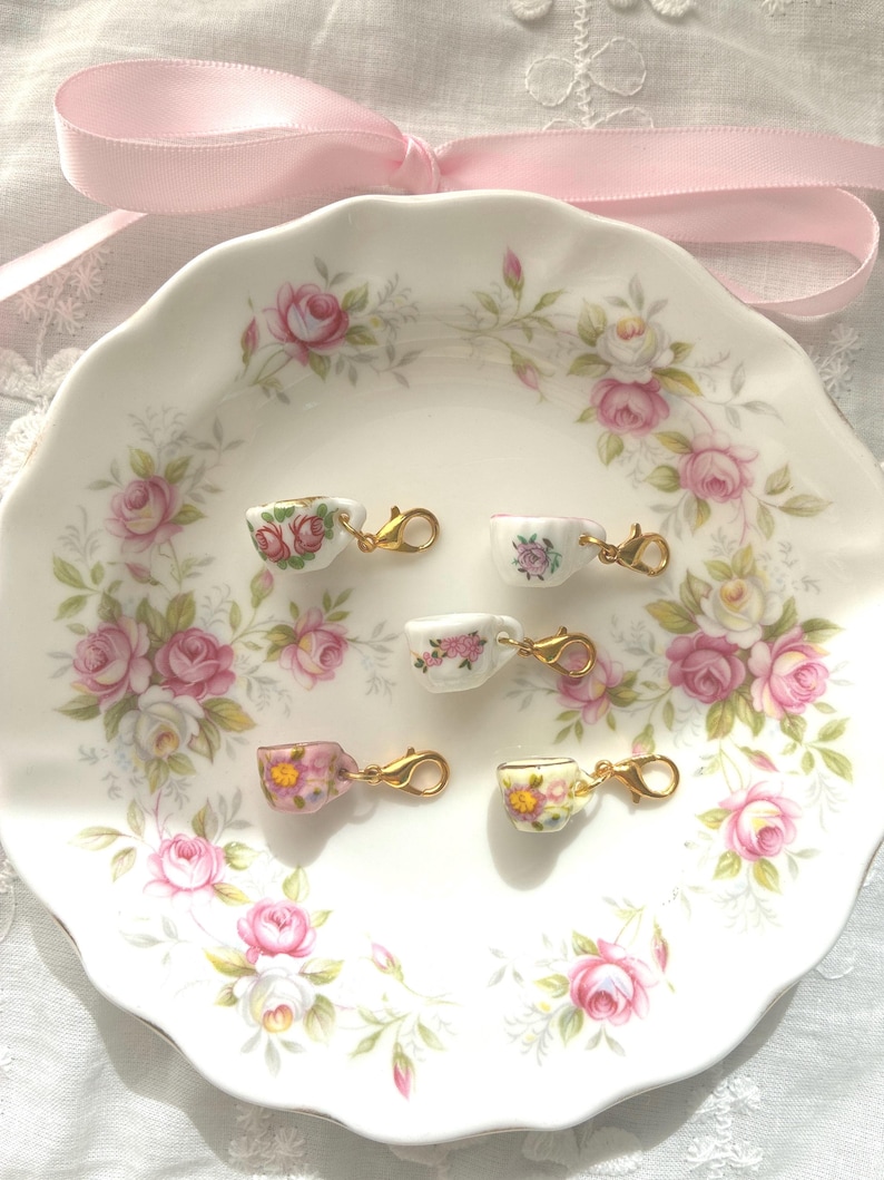 May include: A white ceramic plate with a floral rose pattern holds five miniature teacups with gold-colored clasps. Each teacup features a different floral design. A pink ribbon is tied in a bow above the plate.