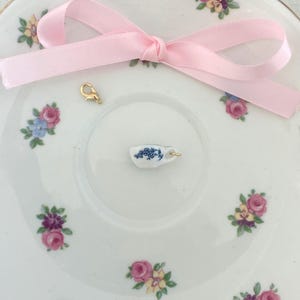 May include: A white porcelain plate with floral designs and a miniature teacup charm. A pink ribbon is tied in a bow, and a gold-colored clasp is also present. The teacup charm has a blue floral pattern.