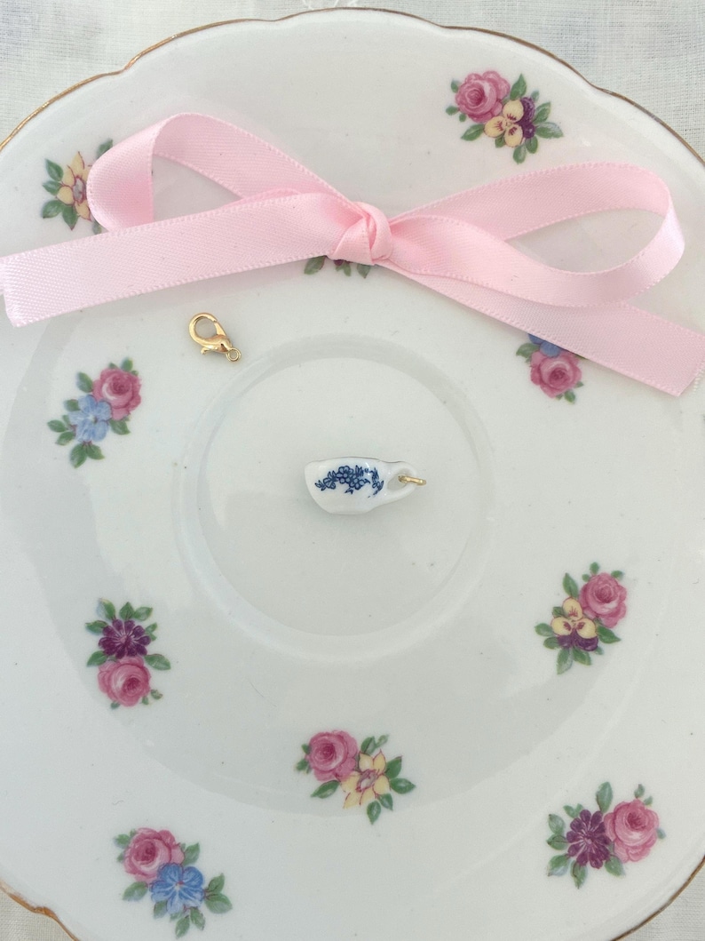 May include: A white porcelain plate with floral designs and a miniature teacup charm. A pink ribbon is tied in a bow, and a gold-colored clasp is also present. The teacup charm has a blue floral pattern.