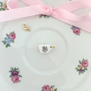 May include: A miniature porcelain teacup charm with a gold-colored handle, featuring a pink rose design. The charm sits on a white saucer decorated with floral patterns. A pink ribbon and a gold clasp are also present.