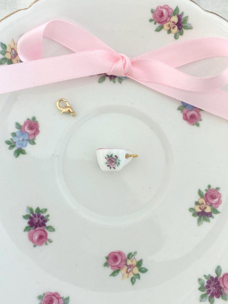 May include: A miniature porcelain teacup charm with a gold-colored handle, featuring a pink rose design. The charm sits on a white saucer decorated with floral patterns. A pink ribbon and a gold clasp are also present.