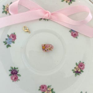 May include: A miniature pink teacup charm with floral details, resting on a white saucer decorated with pink, purple, and blue floral patterns. A light pink ribbon is tied in a bow, and a gold-colored clasp is also present.