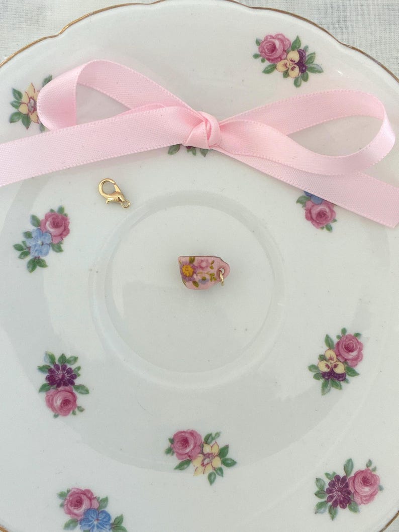 May include: A miniature pink teacup charm with floral details, resting on a white saucer decorated with pink, purple, and blue floral patterns. A light pink ribbon is tied in a bow, and a gold-colored clasp is also present.