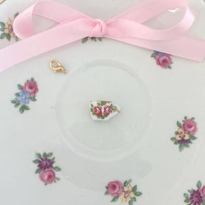 May include: A delicate white porcelain saucer with floral patterns and a pink ribbon bow. A tiny teacup charm with red roses and a gold clasp are on the saucer. The saucer has a gold rim.