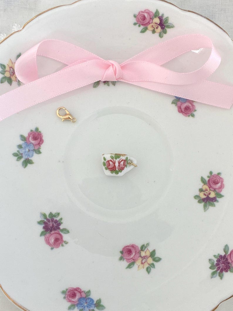 May include: A delicate white porcelain saucer with floral patterns and a pink ribbon bow. A tiny teacup charm with red roses and a gold clasp are on the saucer. The saucer has a gold rim.