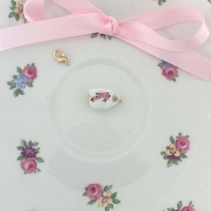 May include: A miniature white teacup charm with pink floral details and a gold-colored clasp sits on a vintage-style saucer. A pink satin ribbon is tied in a bow. The saucer is decorated with pink, purple, and blue floral patterns.
