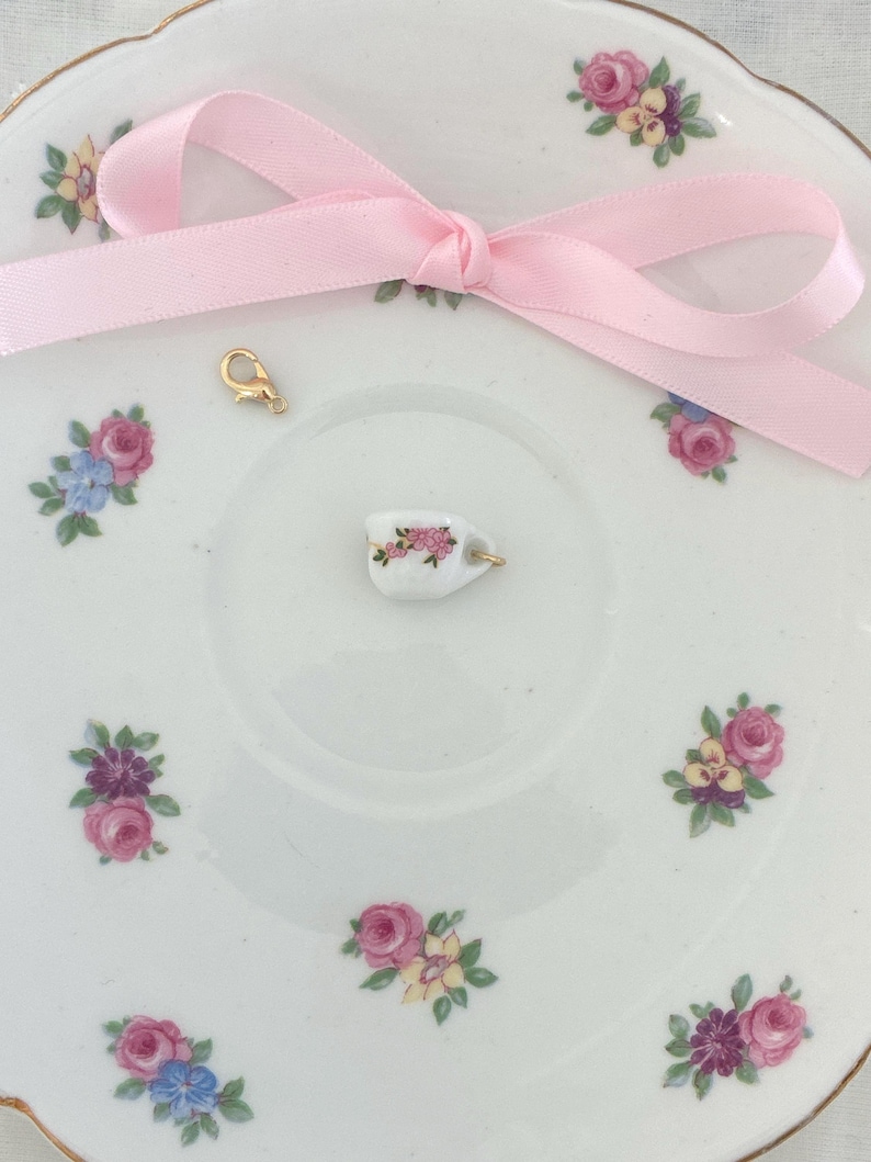 May include: A miniature white teacup charm with pink floral details and a gold-colored clasp sits on a vintage-style saucer. A pink satin ribbon is tied in a bow. The saucer is decorated with pink, purple, and blue floral patterns.