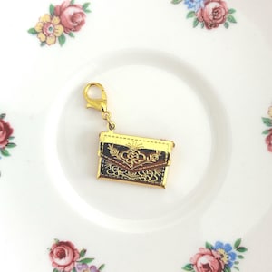 May include: A gold-toned metal charm shaped like a small envelope or wallet, featuring a dark brown rectangular body with an embossed floral pattern on the flap and a gold lobster clasp attached to the top for easy fastening to jewellery or accessories.