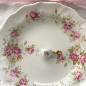 May include: A white ceramic plate with a pink rose pattern, holding a miniature teacup charm. The charm is white with a pink rose design and a gold-colored clasp. A pink ribbon is tied in a bow in the background.