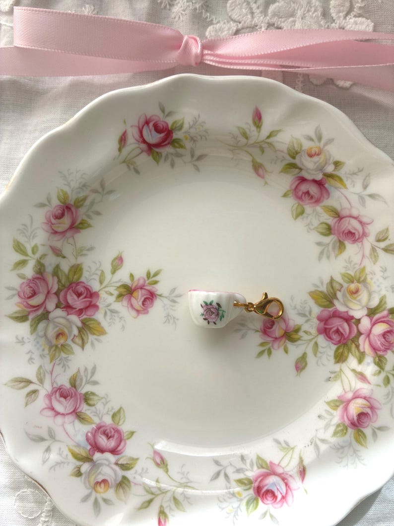 May include: A white ceramic plate with a pink rose pattern, holding a miniature teacup charm. The charm is white with a pink rose design and a gold-colored clasp. A pink ribbon is tied in a bow in the background.