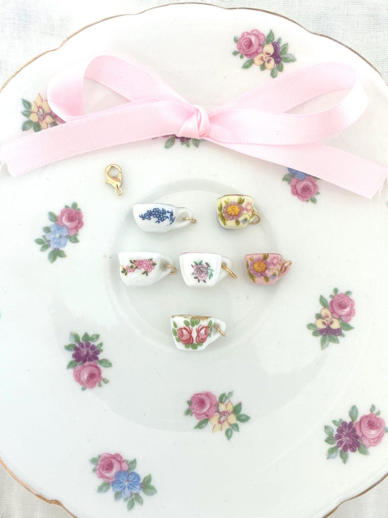 May include: Miniature porcelain teacups with floral patterns, displayed on a white plate adorned with pink and purple roses. A pink ribbon and gold clasp are also present.