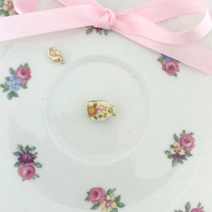 May include: A miniature teacup charm with floral designs, resting on a white saucer decorated with pink and purple roses and blue flowers. A gold-colored clasp and a pink ribbon bow are also present.