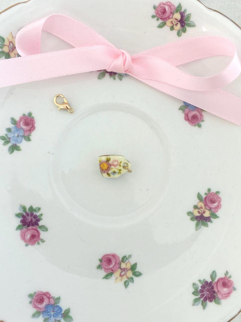 May include: A miniature teacup charm with floral designs, resting on a white saucer decorated with pink and purple roses and blue flowers. A gold-colored clasp and a pink ribbon bow are also present.
