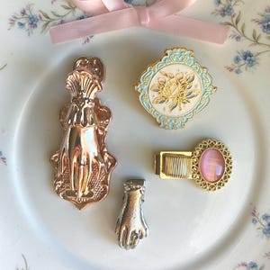 May include: A collection of decorative brooches and pins, featuring a hand-shaped pin, a floral design, and a hair clip with a pink stone. A pink ribbon bow sits above. The items are displayed on a floral patterned plate.